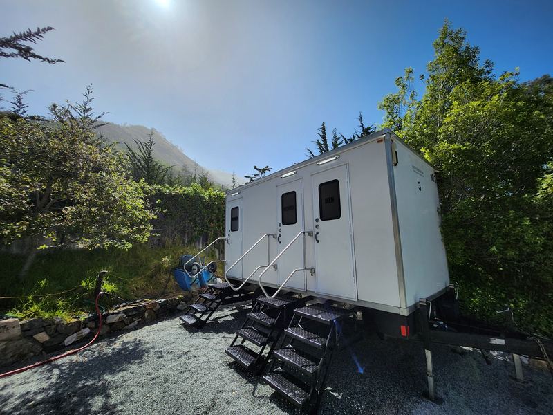 Three-stall restroom trailer on gravel beside green hedges