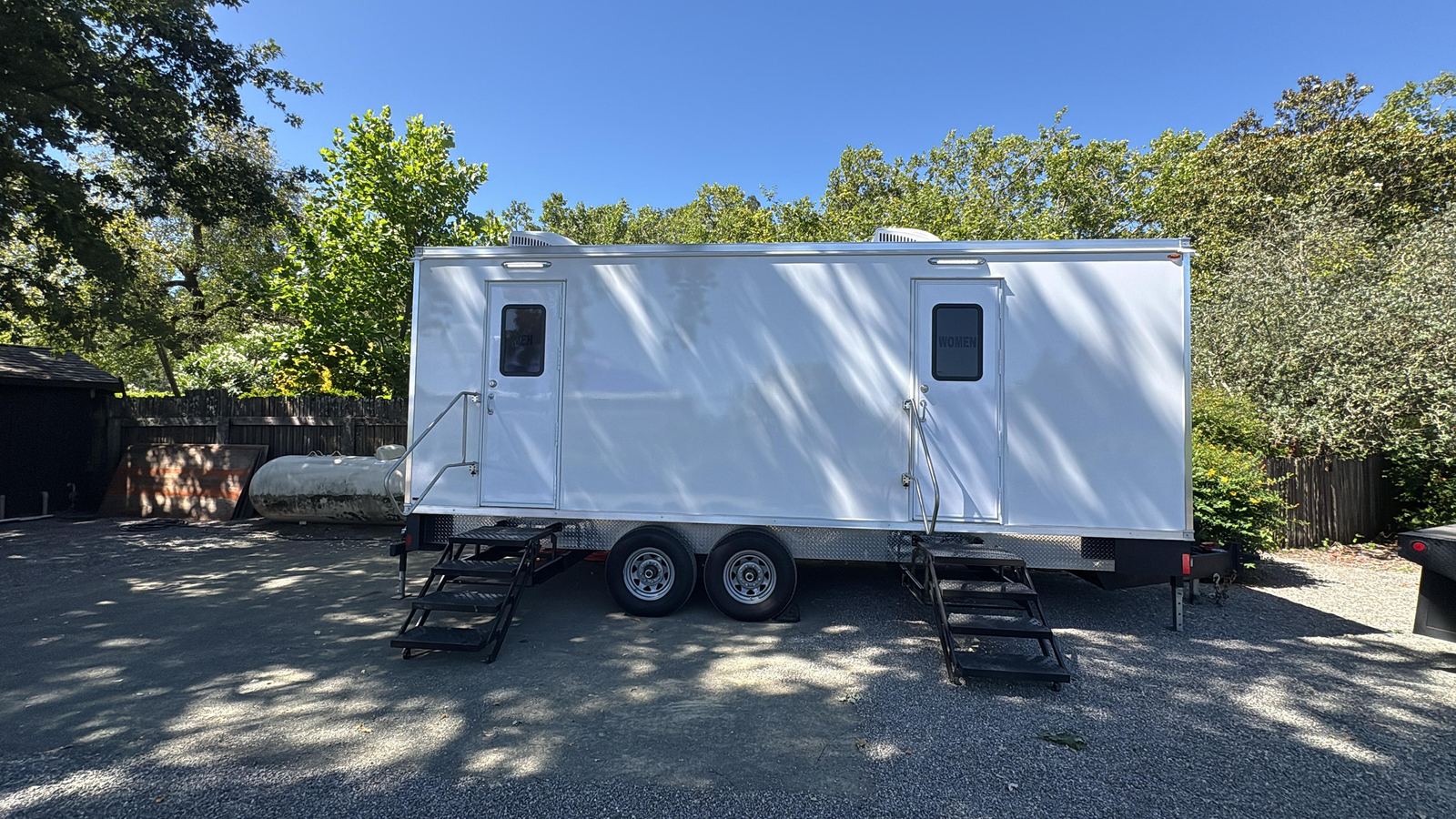 Two-room portable restroom trailer parked in the shade of tall trees