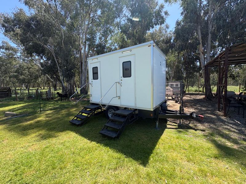 Restroom trailer on grass under shade trees at a park event